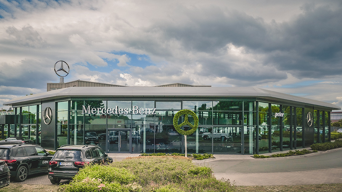 Außenansicht eines modernen Mercedes-Benz Autohauses Peter in Nordhausen mit großflächiger Glasfassade, Mercedes-Benz Logos auf dem Dach und als Heckenschnitt, mehrere schwarze Fahrzeuge im Vordergrund, bewölkter Himmel.