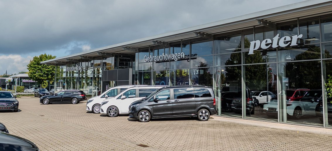 Autohaus Peter mit gro&szlig;er Glasfassade und der Aufschrift �Gebrauchtwagen�. Davor stehen zahlreiche Mercedes-Fahrzeuge, &uuml;berwiegend schwarze und wei&szlig;e Modelle. Links im Hintergrund ist das Mercedes-Benz-Logo auf einem Mast sichtbar. 