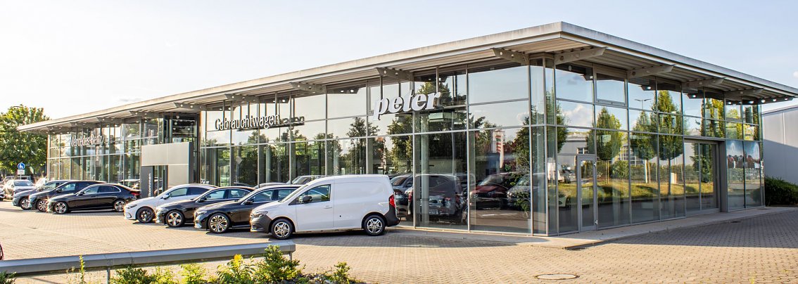 Autohaus Peter mit gro&szlig;er Glasfassade und der Aufschrift �Gebrauchtwagen�. Davor stehen zahlreiche Mercedes-Fahrzeuge, &uuml;berwiegend schwarze und wei&szlig;e Modelle. Links im Hintergrund ist das Mercedes-Benz-Logo auf einem Mast sichtbar. 