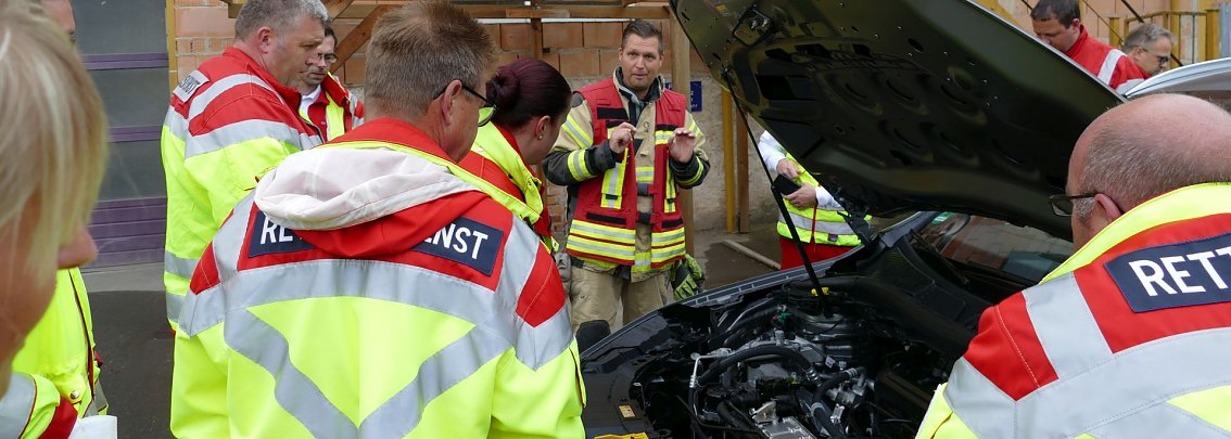 Sandro Hertel (Mitte) von der Berufsfeuerwehr erkl&auml;rt den Rettungskr&auml;ften des DRK, worauf sie im Fall eines verunfallten Elektrofahrzeuges achten m&uuml;ssen. Die gr&ouml;&szlig;te Gefahr geht von der Hochvolt-Anlage aus. Deshalb muss sie als erstes deaktiviert werden.