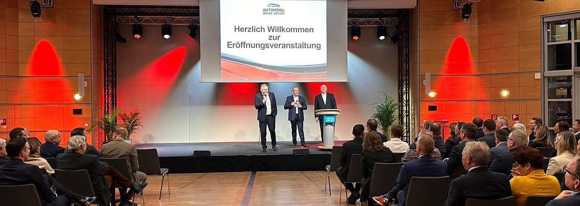 Auf einer gro&szlig;en Leinwand steht Die Automobilmesse Erfurt 2025 ist er&ouml;ffnet!. Davor sprechen drei M&auml;nner (rechts Helmut Peter) auf einer B&uuml;hne, w&auml;hrend zahlreiche Besucher im Saal zuh&ouml;ren.