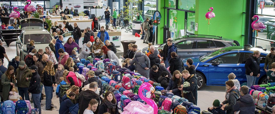 Gro&szlig;er Ausstellungsraum im �koda Autohaus, gef&uuml;llt mit vielen Menschen. In der Mitte sind lange Tische voller bunter Schulranzen. Mehrere �koda-Fahrzeuge stehen zwischen den Besuchern. Rosa Flamingo-Dekorationen h&auml;ngen im Raum.