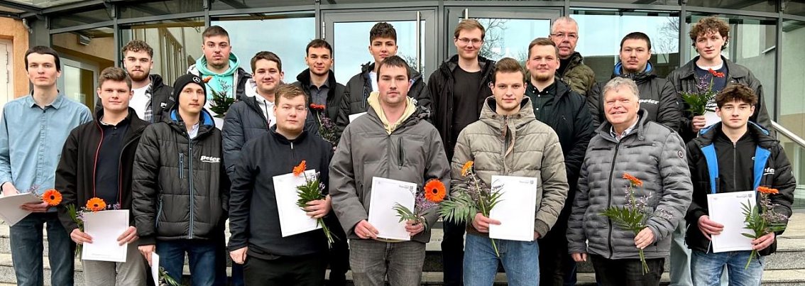 16 junge M&auml;nner der Autohaus Peter Gruppe stehen mit ihren Abschlusszeugnissen und Blumen zusammen mit einem Ausbildungsleiter und einem Berufsschullehrer auf der Treppe vor dem Geb&auml;ude.