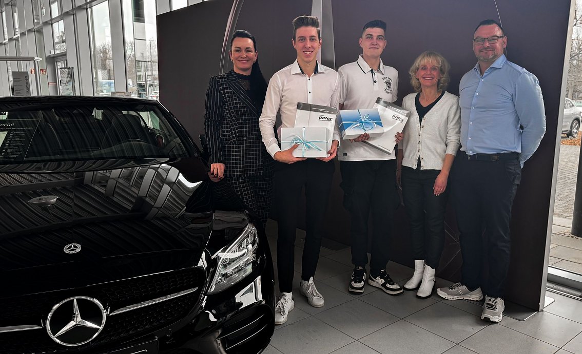 Christina Dost, Gesch&auml;ftsf&uuml;hrerin, steht links neben Paul W. (zweiter von links), der nach verk&uuml;rzter Ausbildung als Assistent der Gesch&auml;ftsleitung startet, und Domenik B. (Mitte), der das Team k&uuml;nftig als Karosseriebauer verst&auml;rkt. Rechts daneben stehen Silke Lang, Ausbildungsleiterin, und Enrico Dost, Serviceleiter. Paul W. und Domenik B. halten jeweils eine Geschenkbox mit hellblauer Schleife in den H&auml;nden. Die Gruppe steht im Autohaus vor einem schwarzen Auto und einer Wanddekoration und l&auml;chelt in die Kamera.