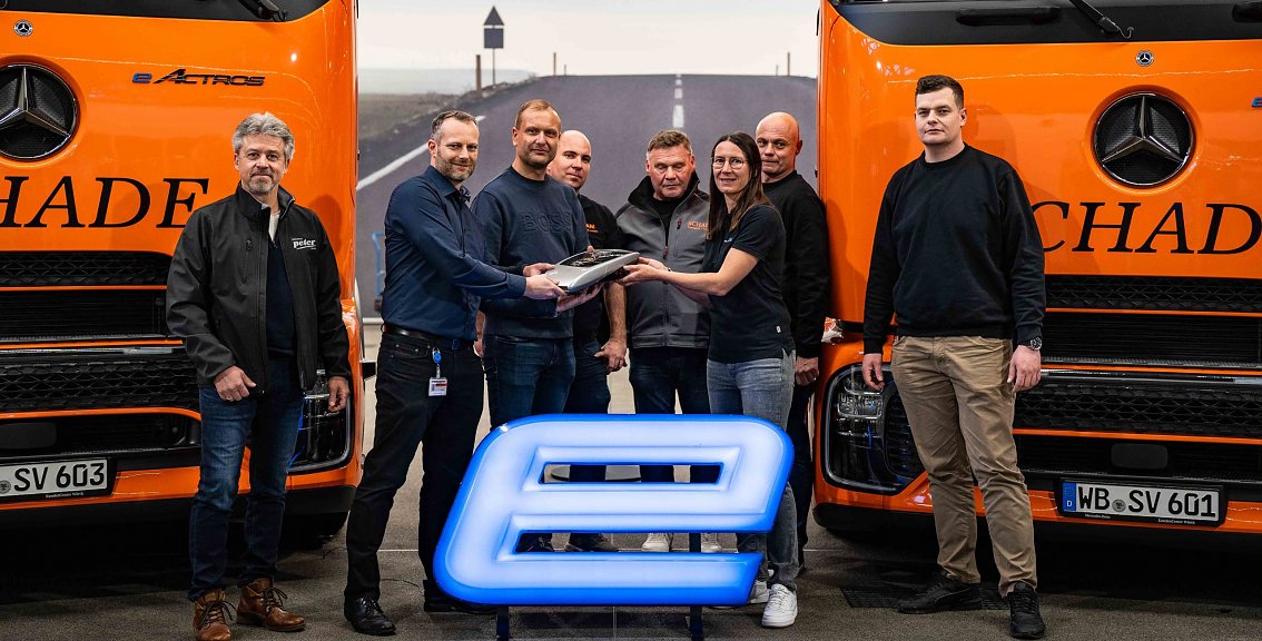 Mehrere Personen vom Autohaus Peter und von Schade Logistics bei der symbolischen &Uuml;bergabe eines Lkw-Schl&uuml;ssels vor zwei orangefarbenen Mercedes-Benz-Lkw. Leuchtendes blaues E-Symbol im Vordergrund.