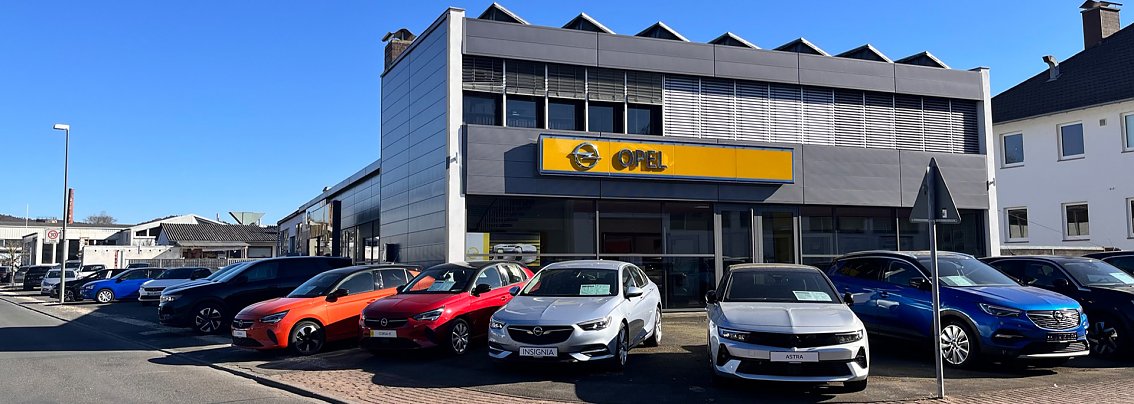 Außenansicht des Opel-Autohauses in Witzenhausen mit mehreren bunten Fahrzeugen (u.a. rot, orange, blau, silber) auf dem Parkplatz vor dem Gebäude. Über dem Eingang ist ein großes gelbes Opel-Logo zu sehen. Der Himmel ist blau und die Sonne scheint.