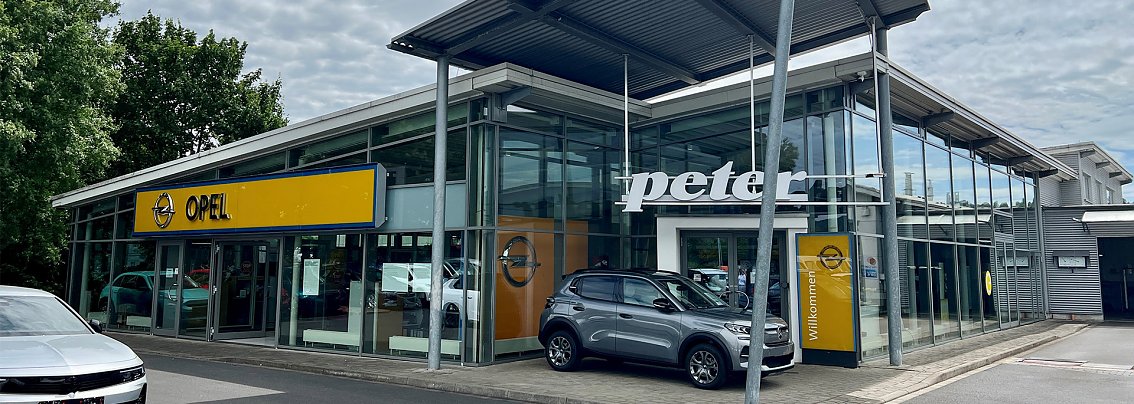 Außenansicht des Autohauses am Standort Eschwege bei sonnigem Wetter. Im Vordergrund zahlreiche parkende Fahrzeuge, im Hintergrund ein modernes Gebäude mit großer Glasfassade und gelbem Opel-Logo.