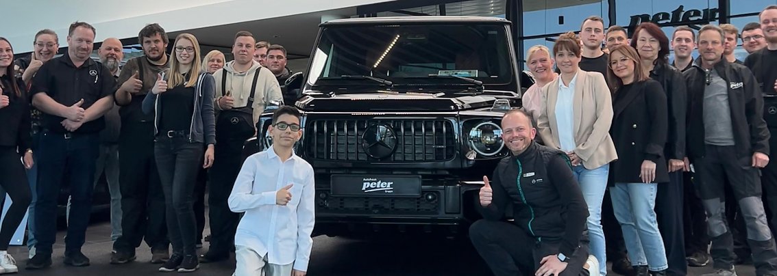 Jamil aus der TV-Show �The Voice Kids� steht mit einem Daumen-hoch-Zeichen vor einem schwarzen Mercedes-Gel&auml;ndewagen im Autohaus Mercedes Wittenberg, umgeben von Mitarbeiterinnen und Mitarbeitern des Autohauses.