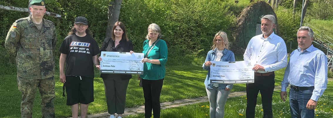 Gruppenfoto bei Spenden&uuml;bergabe zum 24-Stunden-Schwimmen: Sieben Personen stehen im Freien auf einer Wiese, darunter ein Soldat in Uniform, Teilnehmende in Freizeitkleidung und Vertreter des Autohauses Peter. Zwei gro&szlig;e Spendenschecks werden in die Kamera gehalten. Die Stimmung wirkt freundlich und gemeinschaftlich.