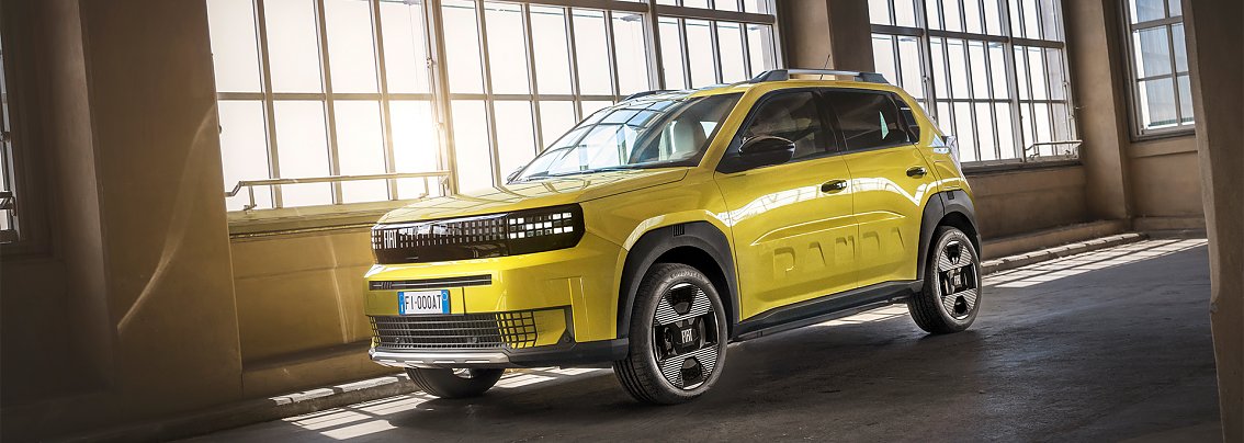 Ein leuchtend gelber Fiat Grande Panda steht schr&auml;g von vorn fotografiert in einer lichtdurchfluteten Industriehalle. Das kompakte SUV im B-Segment hat markante schwarze Akzente an Front, Radk&auml;sten und Dachreling. Auf der Seite ist dezent der Schriftzug PANDA eingearbeitet. Sonnenlicht f&auml;llt durch hohe Fenster und spiegelt sich auf der gl&auml;nzenden Karosserie.