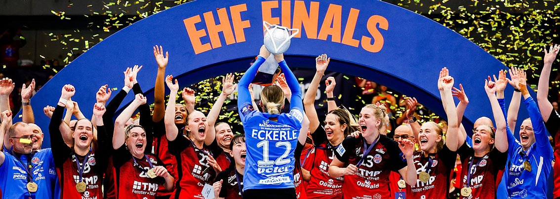Das Bild zeigt ein Handballteam, das gerade den Sieg bei den EHF Finals feiert. In der Mitte hebt die Torh&uuml;terin mit der R&uuml;ckennummer 12 (Eckerle) den Pokal &uuml;ber den Kopf, w&auml;hrend gelbe Konfettistreifen durch die Luft wirbeln. Ihre Mitspielerinnen und das Trainerteam jubeln, klatschen und rei&szlig;en die Arme in die H&ouml;he. Alle tragen Medaillen und schwarze-rote Trikots, einige blaue Torwarttrikots. Im Hintergrund ist ein gro&szlig;er blauer Torbogen mit der Aufschrift EHF FINALS zu sehen.