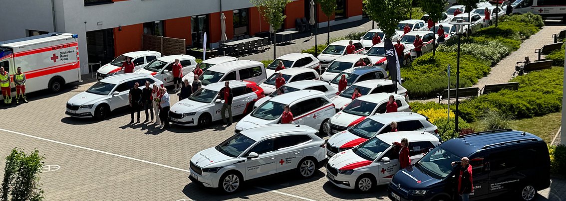 Gruppenfoto mit Fahrzeugflotte: Mehrere neue und bestehende Fahrzeuge des DRK Nordhausen, &uuml;berwiegend wei&szlig; mit rotem Kreuz, stehen in einem Innenhof. DRK-Mitarbeitende in roten Shirts posieren neben den Autos, im Vordergrund ein dunkelblauer Transporter.