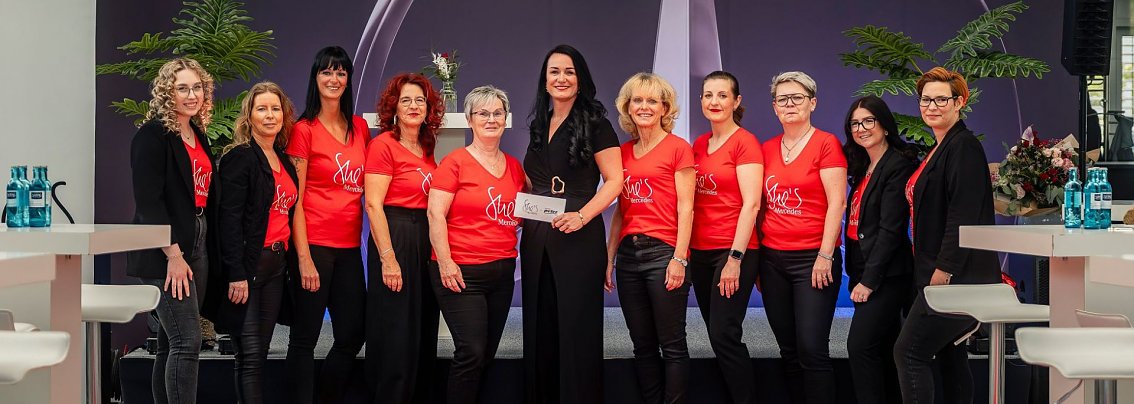 Eine Gruppe von elf Frauen, einige in schwarzen Outfits, andere in roten T-Shirts mit der Aufschrift She�s Mercedes, steht l&auml;chelnd nebeneinander auf einer B&uuml;hne vor einer Wand mit gro&szlig;em Mercedes-Logo.