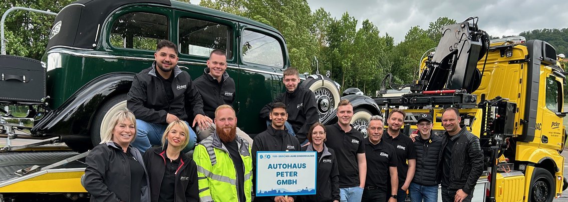 Gruppenfoto eines Teams des Autohaus Peter beim Festumzug in Sondershausen. Die Mitarbeitenden stehen vor einem klassischen Abschleppwagen des ADAC mit einem Oldtimer auf der Ladefl&auml;che. Alle tragen schwarze Autohaus-Peter-Bekleidung, einige lachen in die Kamera. Im Vordergrund wird ein Schild mit der Aufschrift Autohaus Peter GmbH gehalten. Im Hintergrund B&auml;ume und bew&ouml;lkter Himmel.