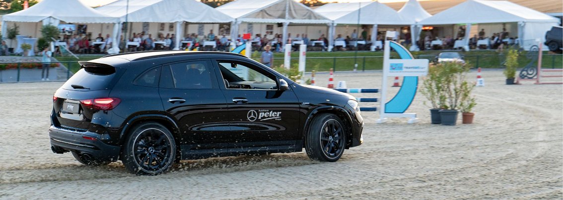 Schwarzer Mercedes-Benz GLA mit Autohaus-Peter-Logo f&auml;hrt beim Jump & Drive &uuml;ber den Reitplatz in Immenrode, im Hintergrund Zuschauer unter wei&szlig;en Pagodenzelten. [Mercedes-AMG GLA 35 4MATIC | Energieverbrauch kombiniert: 9,3-8,8 l/100 km | CO2-Emissionen kombiniert: 211-200 g/km | CO2-Klasse: G]