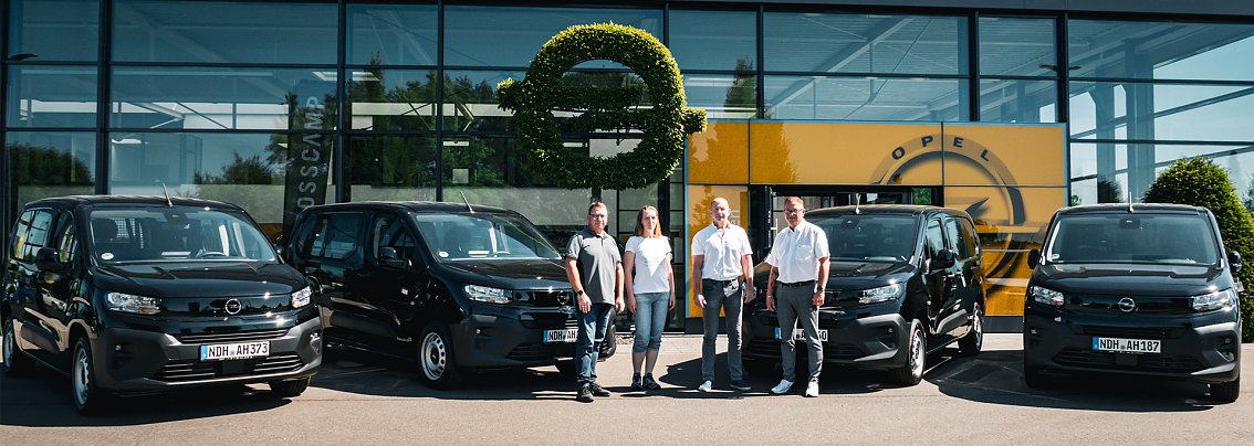 Vor einem modernen Autohaus mit dem Namen Peter stehen vier schwarze Opel-Transporter in einer Reihe. Vier Personen � drei M&auml;nner und eine Frau � posieren jeweils neben den Fahrzeugen. An der Glasfassade ist das Opel-Logo und ein gelber Hintergrund zu sehen.