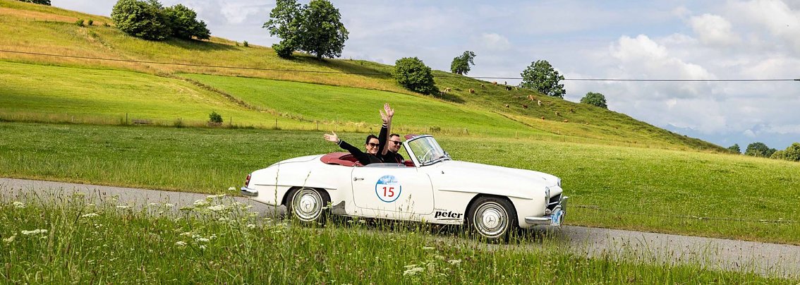Christina und Enrico Dost winken fr&ouml;hlich aus einem wei&szlig;en Mercedes-Benz 190 SL, einem offenen Oldtimer-Cabriolet (Rennnummer 15) auf einer schmalen Landstra&szlig;e, im Hintergrund saftig-gr&uuml;ne Wiesen, sanfte H&uuml;gel und vereinzelt B&auml;ume unter leicht bew&ouml;lktem Himmel.