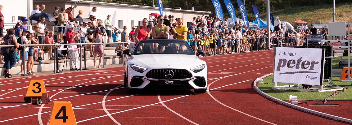 Wei&szlig;es Mercedes-Cabrio f&auml;hrt auf die rote Tartanbahn des Stadions ein. Auf dem R&uuml;cksitz sitzen mehrere Sportlerinnen, w&auml;hrend Zuschauer am Rand applaudieren.