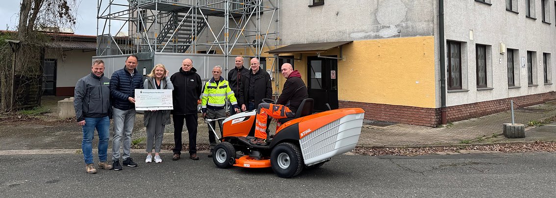 Mehrere Personen vor einem Geb&auml;ude, eine Person sitzt auf einem Rasentraktor, eine weitere h&auml;lt einen gro&szlig;en Scheck.