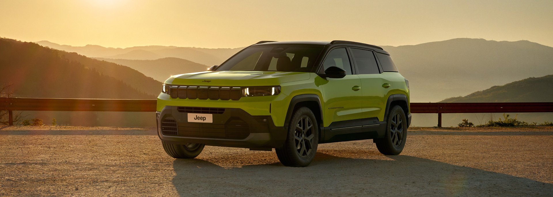 Der neue Jeep&reg; Compass e-Hybrid - Gr&uuml;ner Jeep Compass SUV steht bei Sonnenuntergang auf einem Aussichtspunkt mit Berglandschaft im Hintergrund.