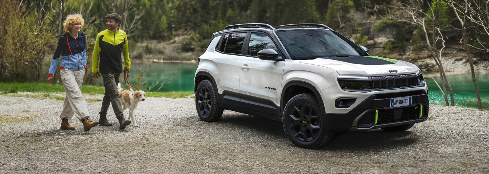 Jeep&reg; Avenger 4xe - Wei&szlig;er Jeep Avenger 4xe steht vor einem See, daneben gehen zwei Menschen in Outdoor-Kleidung mit einem Hund an der Leine spazieren.