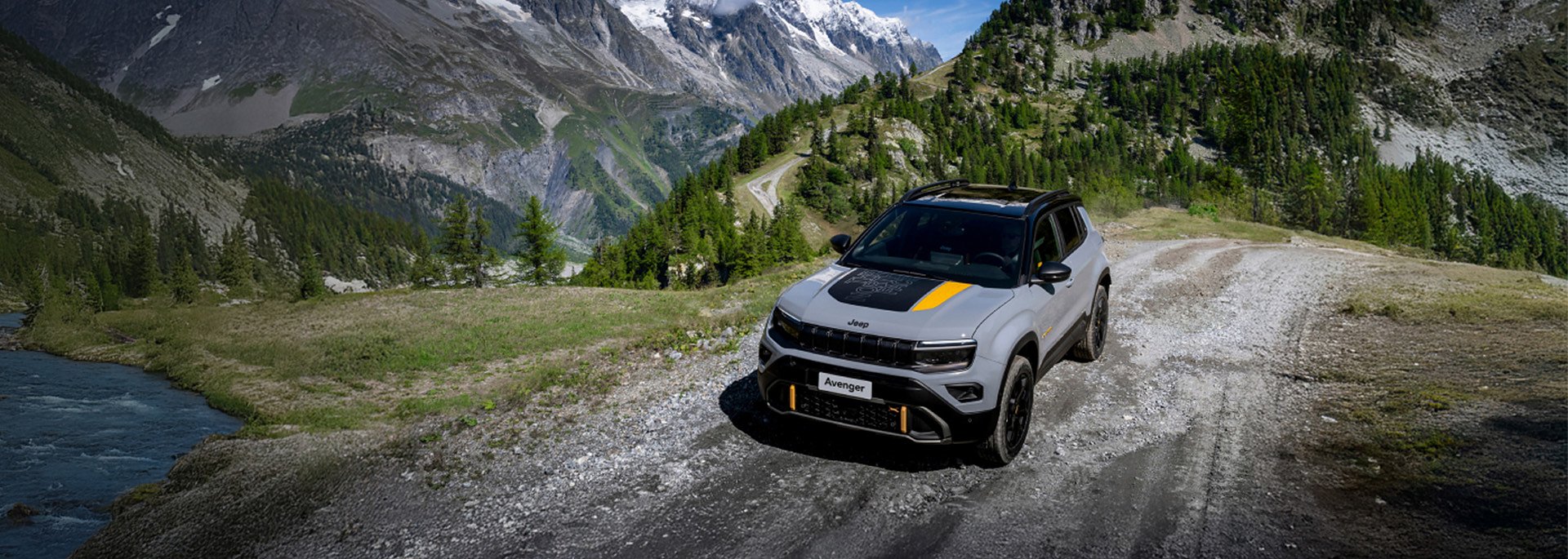 Grauer Jeep Avenger f&auml;hrt auf einem Schotterweg durch eine alpine Berglandschaft mit Fluss, W&auml;ldern und hohen Felsen im Hintergrund.