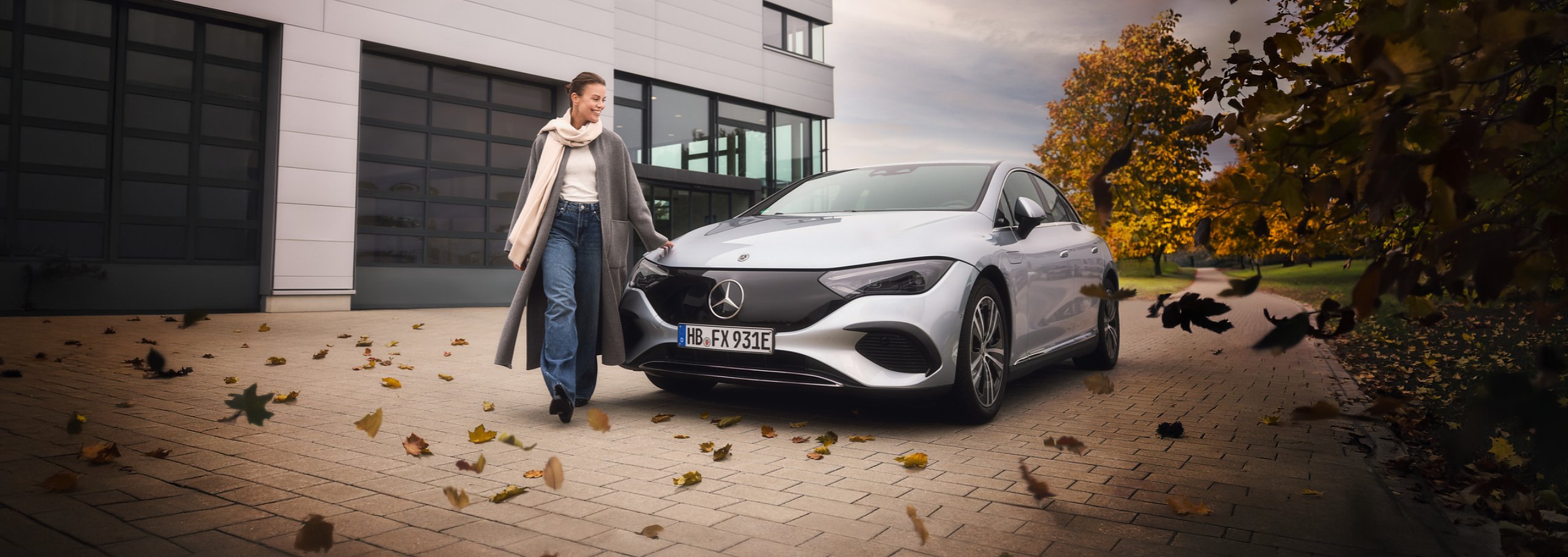 Eine Frau im langen Mantel geht neben einem silbernen Mercedes-Fahrzeug. Auf dem Pflaster liegen bunte Herbstbl&auml;tter, im Hintergrund stehen ein modernes Geb&auml;ude und herbstliche B&auml;ume.