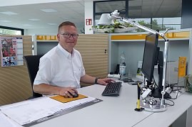 Ronny Peukert, Verkaufsberater, sitzt freundlich lächelnd an einem modernen Schreibtisch im hellen Autohaus-Büro. Er trägt ein weißes Hemd und eine Brille. Auf dem Schreibtisch stehen ein Monitor, Tastatur, Maus, Schreibtischlampe und dekorative Elemente mit Opel-Logo. Im Hintergrund sind Schränke und Büromaterialien zu sehen.