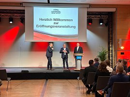 Nahaufnahme der Bühne: Drei Männer im Anzug (rechts Helmut Peter) stehen vor einer Leinwand mit der Begrüßung zur Eröffnungsveranstaltung. Einer spricht ins Mikrofon.