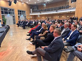 Publikum im Saal hört aufmerksam einer Rednerin auf der Bühne zu. Viele Männer im Anzug, einzelne Frauen im Publikum.