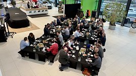 Blick von oben auf den Ausstellungsraum. Mehrere lange Tische sind mit Fr&uuml;hst&uuml;ck gedeckt. Viele Unternehmerinnen und Unternehmer sitzen und diskutieren. Links im Bild ein abgedecktes Fahrzeug.