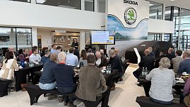 Eine Rednerin steht vor einer Leinwand und pr&auml;sentiert. Auf dem Bildschirm sind Energiediagramme zu sehen. G&auml;ste h&ouml;ren an langen Fr&uuml;hst&uuml;ckstischen aufmerksam zu.