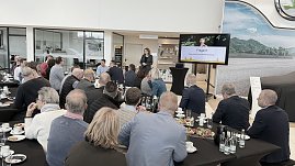 Mehrere Dutzend G&auml;ste sitzen an langen Tischen und schauen auf die Rednerin. Auf dem Bildschirm ist die Abschlussfolie Fragen? zu sehen. Im Vordergrund Teller und Getr&auml;nke.