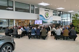 Blick von hinten auf die G&auml;ste beim Unternehmerfr&uuml;hst&uuml;ck. Vorne ein Bildschirm mit Pr&auml;sentation, daneben ein schwarzes, abgedecktes Fahrzeug.