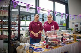 Zwei Frauen stehen hinter einem Kuchen- und Tortenstand im Autohaus. In einer Glasvitrine und auf Tischen sind Cupcakes und Torten ausgestellt. Rosa Flamingos dienen als Dekoration. Ein Banner mit dem Logo Happy Cakes h&auml;ngt &uuml;ber dem Stand.