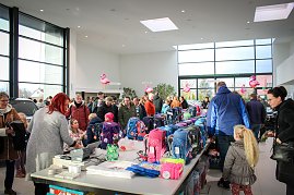 Viele Menschen, darunter Kinder mit Eltern, stehen an Tischen voller bunter Schulranzen im Autohaus. Rosa Flamingo-Dekorationen h&auml;ngen von der Decke. Im Hintergrund sind Autos und gro&szlig;e Fensterfronten sichtbar.