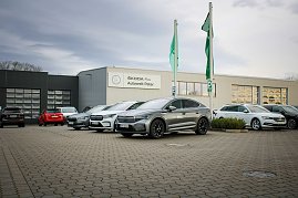 Mehrere �koda-Fahrzeuge stehen vor dem Geb&auml;ude �koda Plus Autowelt Peter. Zwei gr&uuml;ne Fahnen wehen im Vordergrund. Links im Bild parken weitere Autos.