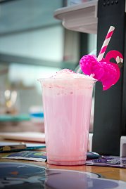 Ein rosa Milchshake oder Cocktail in einem Plastikbecher mit Sahnehaube. Am Strohhalm ist eine pinke Flamingo-Deko befestigt.