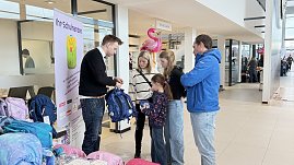Ein Verk&auml;ufer zeigt einer Familie mit zwei Kindern einen blauen Schulranzen. Neben ihnen stehen weitere Ranzen. Im Hintergrund sind Flamingos und Schilder im Autohaus zu sehen.