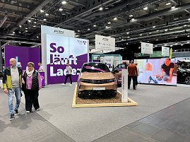 Ein goldfarbener Opel Grandland Electric auf einem Podest mit Holzunterlage, dahinter gro&szlig;e lilafarbene Messewand mit Schriftzug So l&auml;uft Laden.