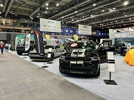 Ein schwarzer Dodge mit gelben Rallyestreifen vorne und ein grauer Dodge Challenger stehen nebeneinander. Daneben schwarze RAM-Fahrzeuge mit gro&szlig;en Bannern.