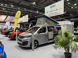 Grauer Opel Crosscamp mit ge&ouml;ffnetem Hochdach und Schiebet&uuml;r, pr&auml;sentiert auf der Messe. Daneben Besucher und weitere Opel-Fahrzeuge.