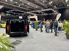Schwarzer Transporter mit ge&ouml;ffneter Heckklappe, ausgestellt auf der Messe. Besucher laufen durch die Halle, viele Fahrzeuge im Hintergrund.