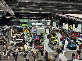 Panoramaaufnahme der Messehalle mit zahlreichen Autos in verschiedenen Farben, vielen Besuchern und gro&szlig;en Markenbannern.