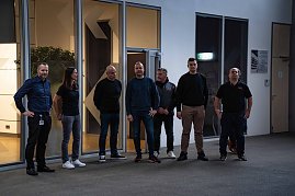 Acht Personen stehen zusammen in einer Halle, im Hintergrund Glasfronten und T&uuml;ren.