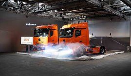 "Zwei orangefarbene Mercedes-Benz eActros-Lkw mit Schade-Logo w&auml;hrend einer Fahrzeug-Enth&uuml;llung mit Nebeleffekten.