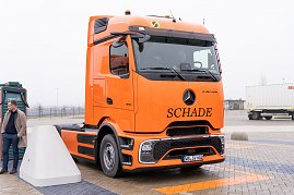 "Orangefarbener Mercedes-Benz eActros mit Schade-Logo auf einem Parkplatz, daneben eine Person und eine Lades&auml;ule.