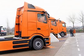 Seitenansicht von drei orangefarbenen Mercedes-Benz eActros 600 Lkw, die in einer Reihe auf einem Hof stehen. Rechts im Bild sind drei Personen im Gespr&auml;ch.