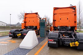 Heckansicht von zwei eActros 600 Lkw beim Laden. Zwischen den Fahrzeugen steht eine gro&szlig;e Lades&auml;ule, im Hintergrund mehrere Personen.