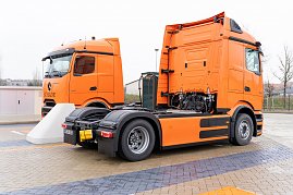 Zwei orangefarbene eActros 600 Lkw stehen nebeneinander an einer Ladestation. Im Hintergrund B&auml;ume und ein Zaun.